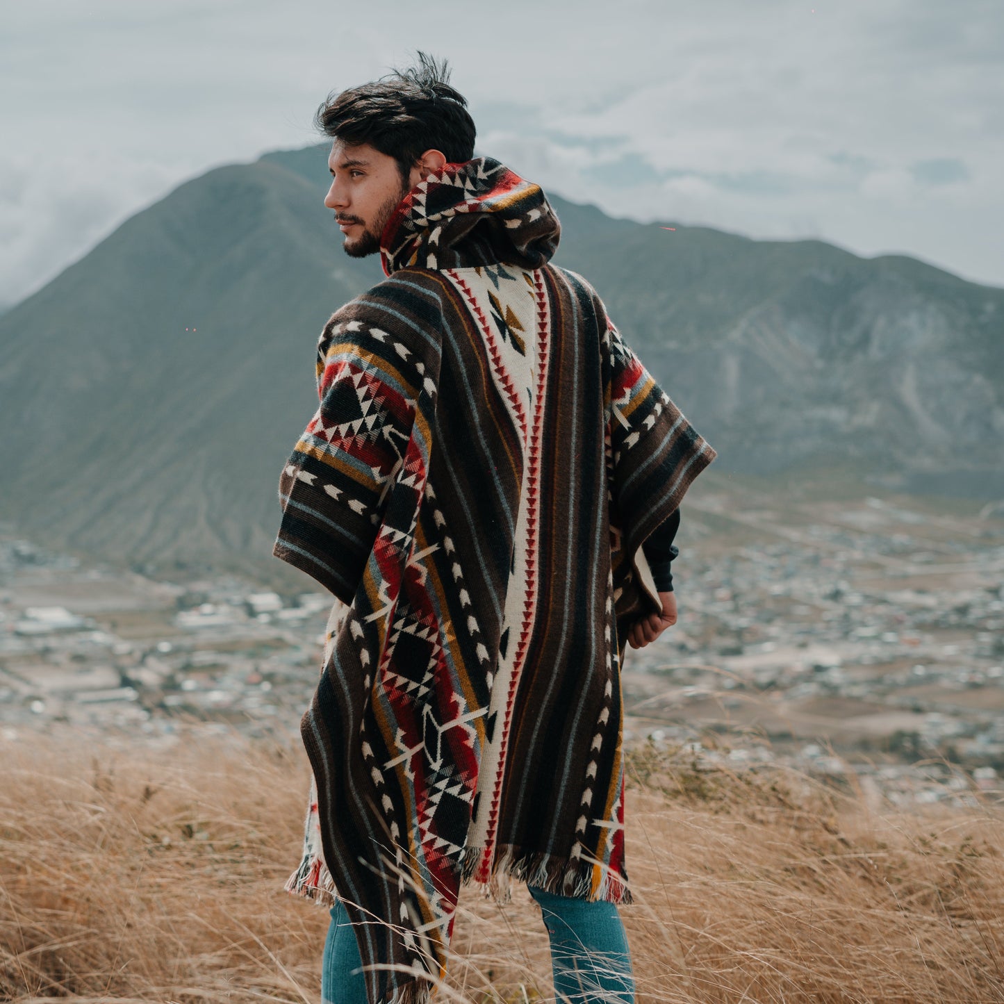 Man wearing a traditional Andean alpaca poncho with hood, multicolor geometric patterns, standing on a mountain in Ecuador overlooking scenic valley, sustainable and ethically crafted fashion.