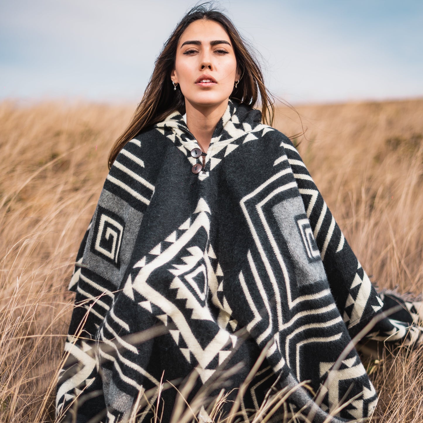Woman wearing black and white geometric alpaca poncho, sitting in tall golden grass under a clear blue sky, looking confidently at the camera.