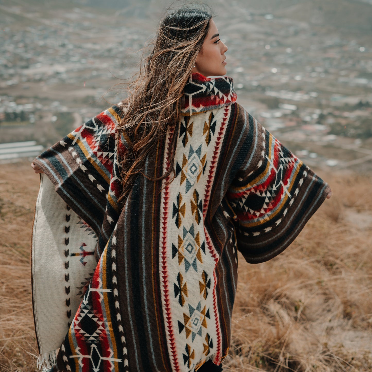 Woman wearing brown and cream alpaca poncho with colorful tribal patterns, standing on a grassy hill with a mountain town in the background, hair flowing in the wind.