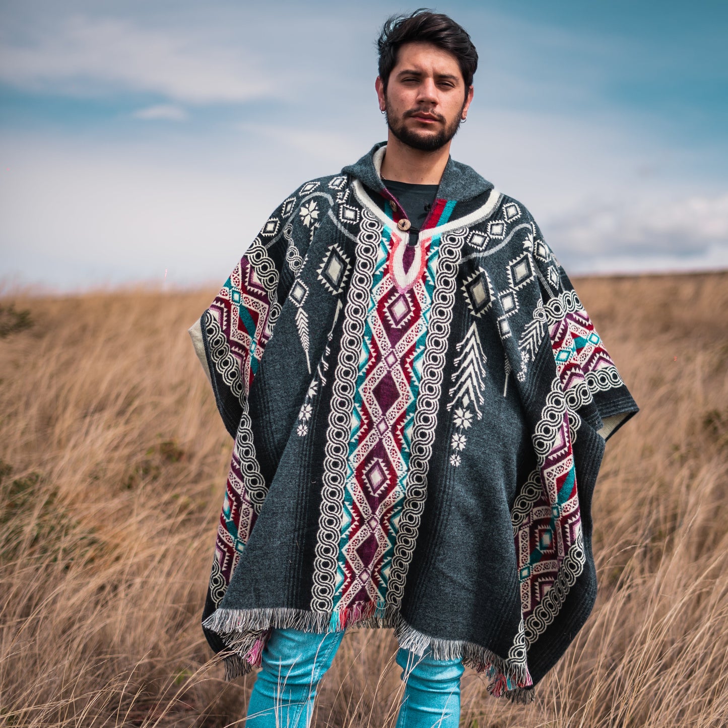 Man wearing a black and burgundy alpaca wool poncho with traditional Andean patterns, handmade in Ecuador by the Otavalo indigenous community, standing in a grassy field.