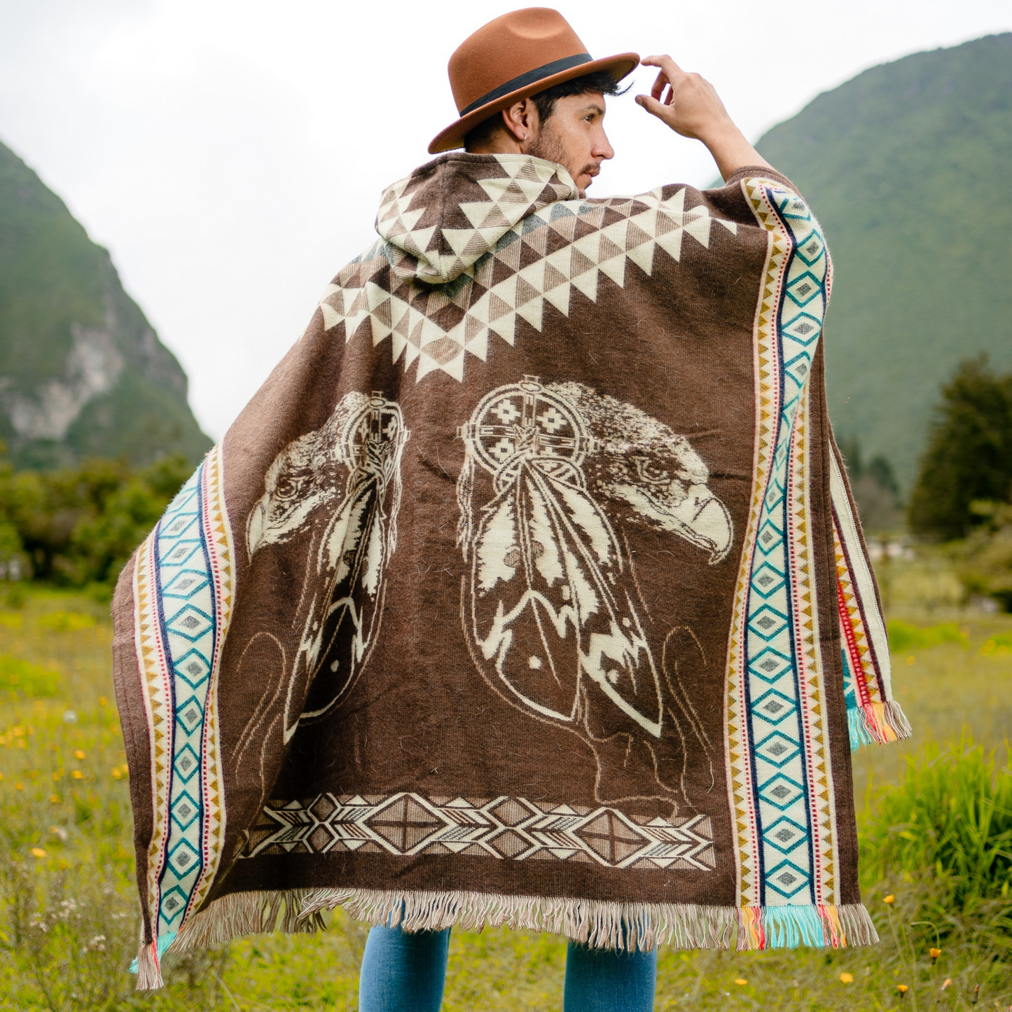 Men’s alpaca wool poncho in rich brown with cream feather and dreamcatcher motifs, accented by turquoise and geometric side borders, featuring a hood and coconut button closure. Handmade by Otavalo indigenous artisans in Ecuador from 70% alpaca wool and 30% recycled orlon, photographed outdoors in the Ecuadorian highlands.