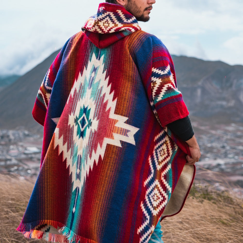 Men’s alpaca wool poncho in vibrant red, blue, and cream tribal pattern with large diamond motif on the back, handmade by Otavalo artisans in Ecuador from 70% alpaca wool and 30% recycled orlon, modeled outdoors in a highland mountain landscape.