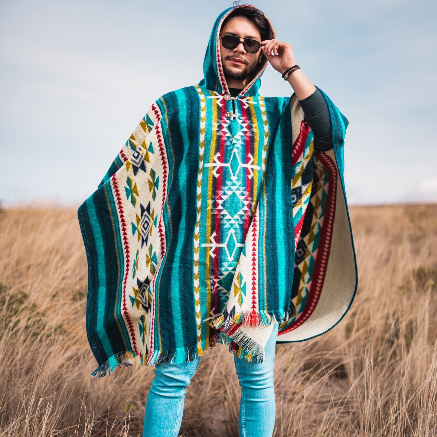 Men’s alpaca wool poncho in teal and white Andean tribal design, handmade in Otavalo, Ecuador, with coconut buttons, crafted from 70% alpaca wool and 30% recycled orlon, modeled outdoors in golden grass fields.