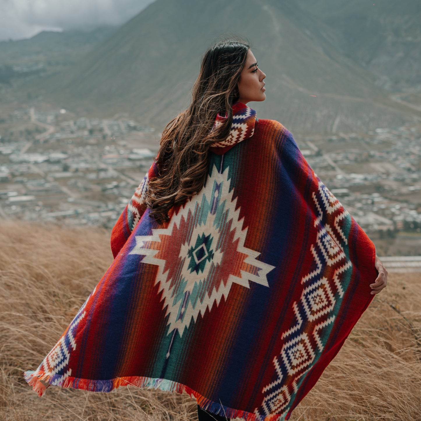 Woman wearing red Andean alpaca poncho with bold geometric diamond patterns, standing on a hillside overlooking a valley town and mountains.
