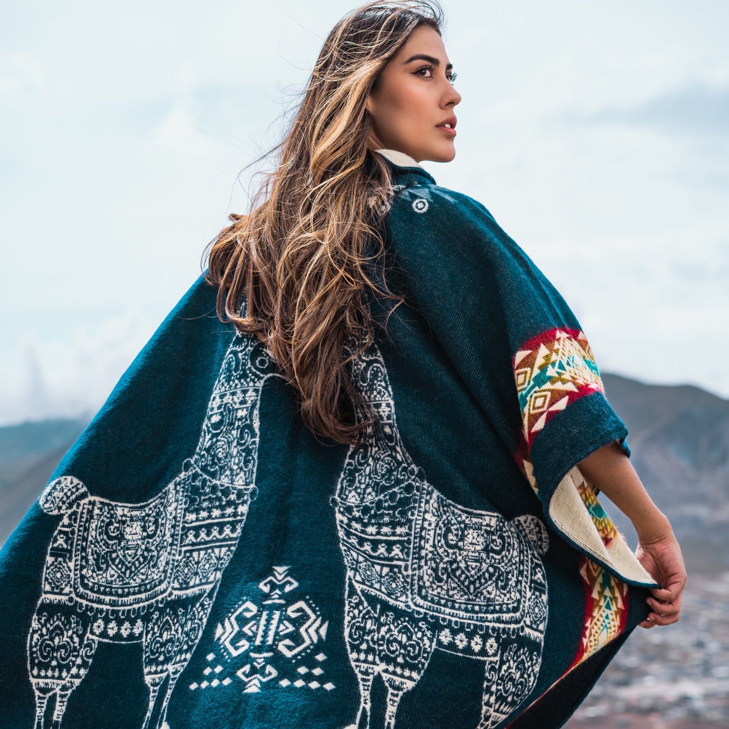 Woman wearing teal alpaca poncho with white llama patterns and colorful Andean stripes, standing on a hillside with a mountain town in the background, hair flowing in the wind.