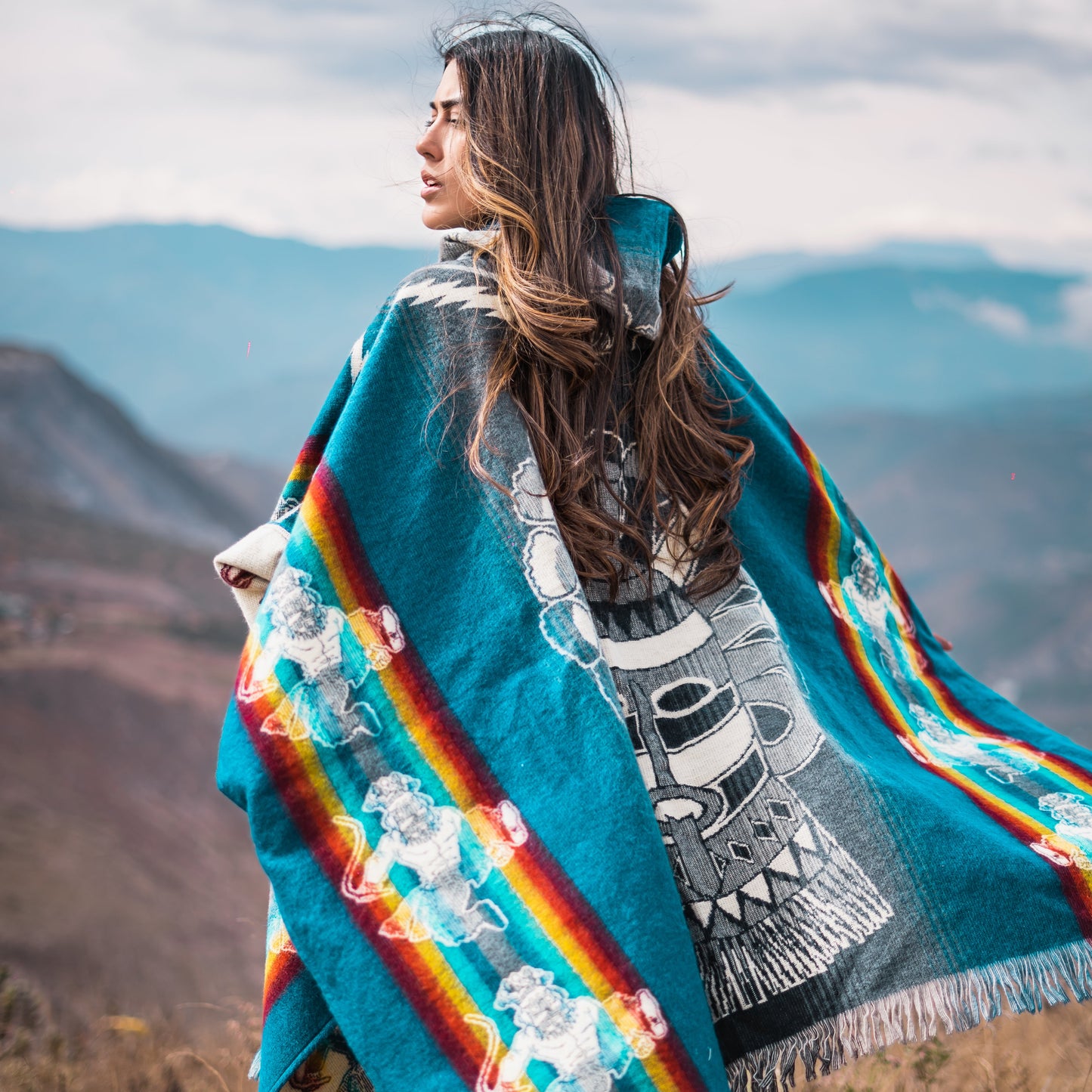 Woman wearing turquoise Andean alpaca poncho with colorful rainbow stripes and warrior motifs, standing on a mountain ridge with dramatic cloudy sky.