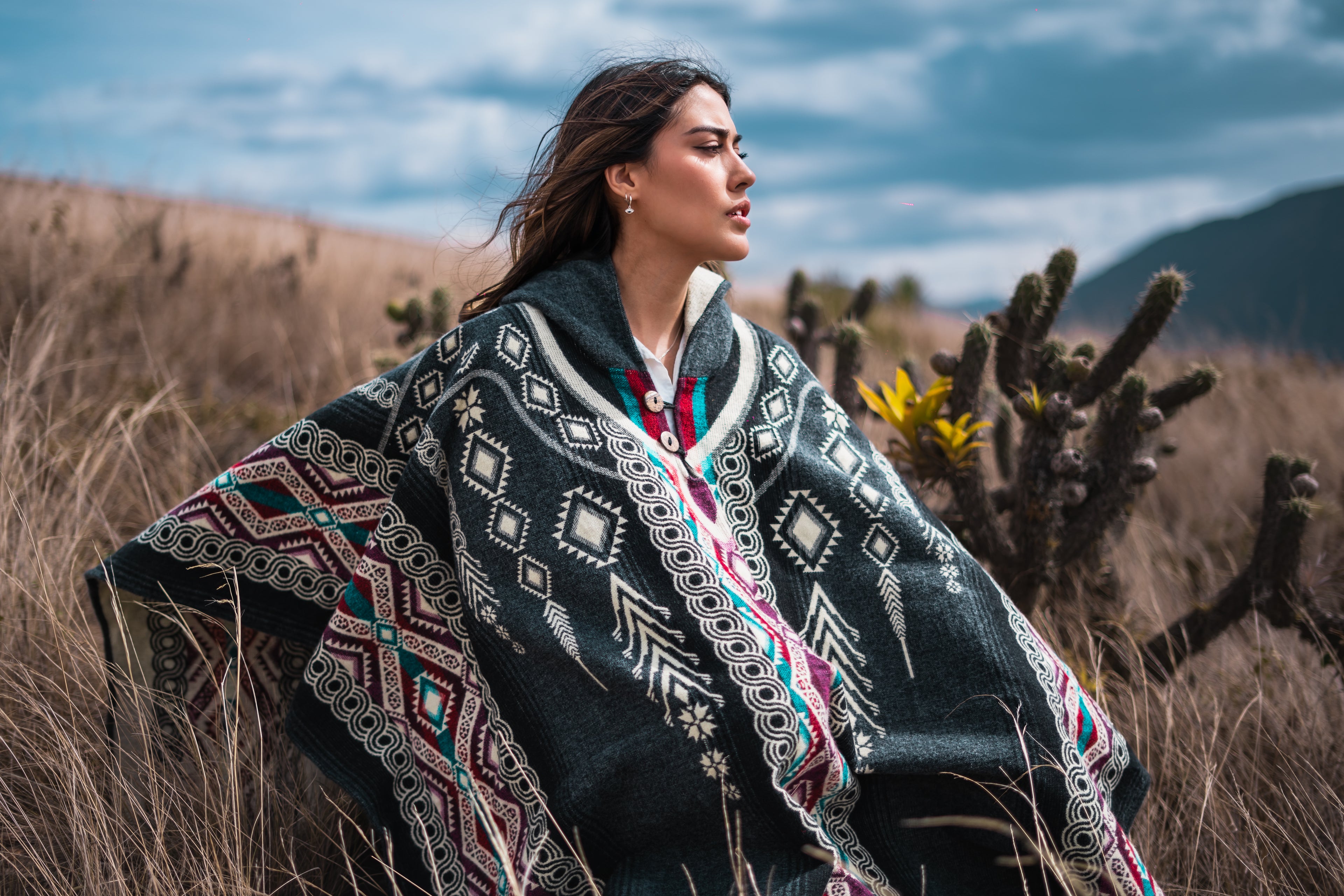 Woman wearing a black alpaca wool poncho with Andean patterns, standing in a desert landscape with cacti and mountains