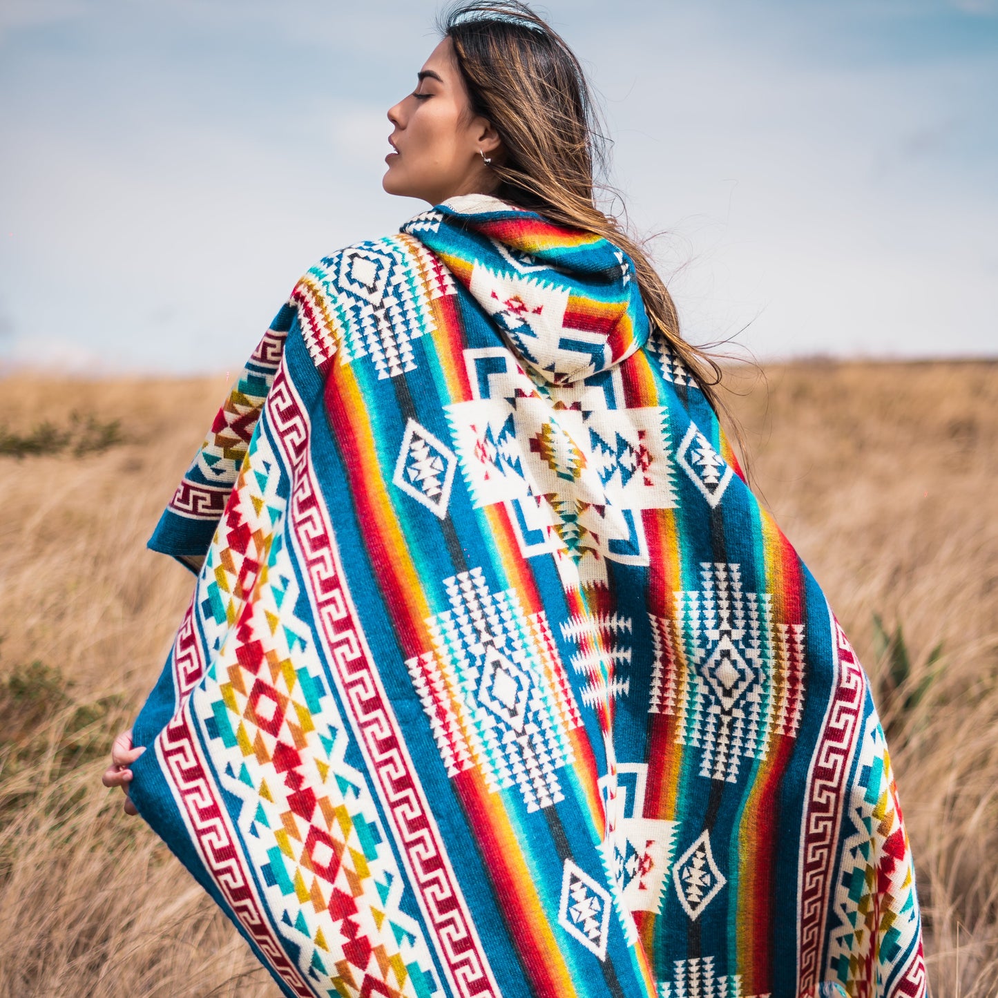 Woman wearing a handmade blue and red rhombus alpaca wool poncho with Andean patterns, standing in a golden grass field under a clear sky.