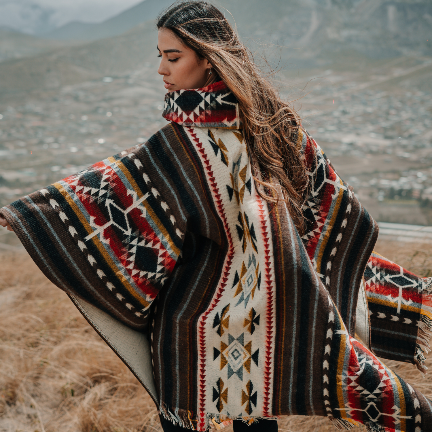 Woman wearing a square-cut alpaca wool poncho with colorful tribal patterns, standing in the Andes mountains with long hair blowing in the wind.