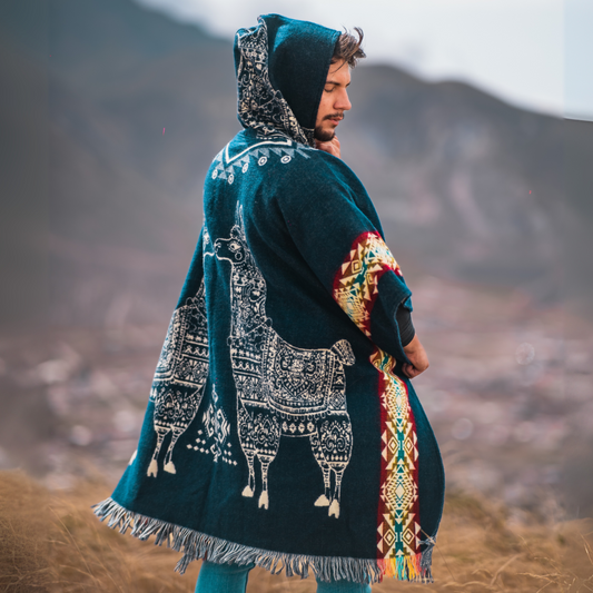 Men’s alpaca wool poncho in deep teal with intricate white llama motifs and multicolor Andean side borders, featuring a hood and fringed hem. Handmade by Otavalo indigenous artisans in Ecuador from 70% alpaca wool and 30% recycled orlon, modeled outdoors in a highland mountain landscape.