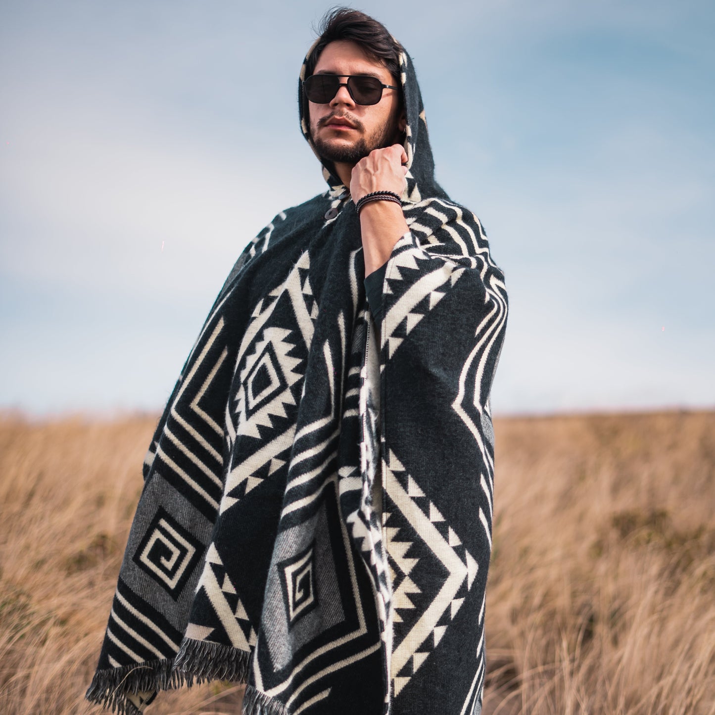 Men’s alpaca wool poncho in teal blue with large Inca mask design, rainbow side accents, and fringed hem, handmade by Otavalo indigenous artisans in Ecuador from 70% alpaca wool and 30% recycled orlon, modeled outdoors in a dry grass highland landscape.