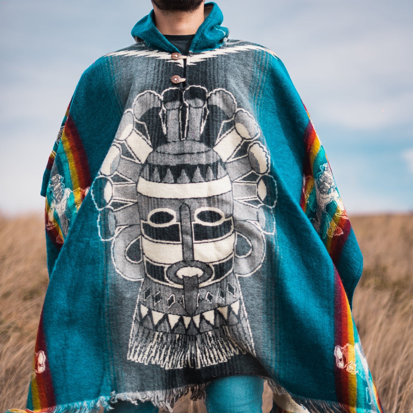 Men’s alpaca wool poncho in teal blue with large Inca mask design, rainbow side accents, and fringed hem, handmade by Otavalo indigenous artisans in Ecuador from 70% alpaca wool and 30% recycled orlon, modeled outdoors in a dry grass highland landscape.