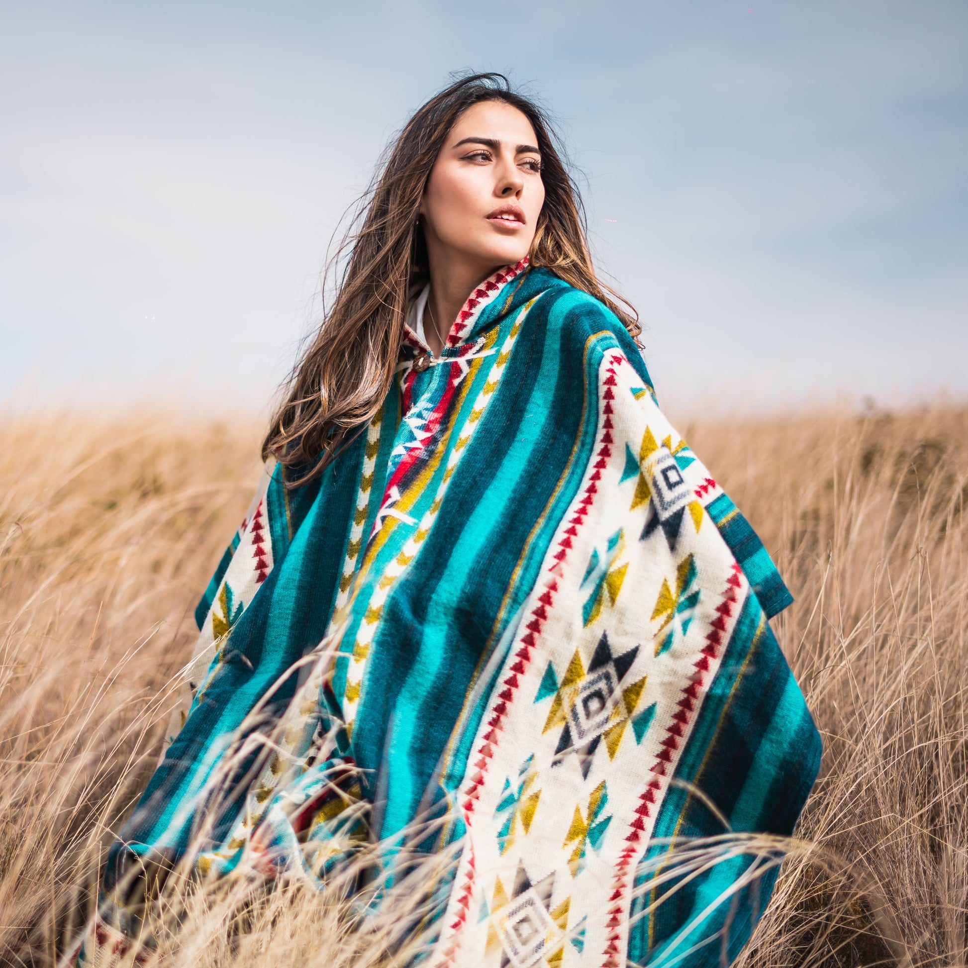 Woman wearing turquoise Andean alpaca poncho with geometric tribal patterns, standing in a golden grass field under a clear sky.